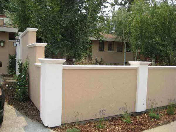 Beige concrete plaster wall with white trim surrounding a house, set in a landscaped area.