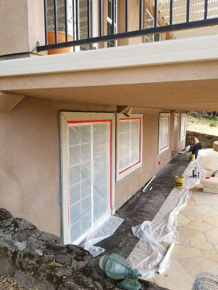 Residential exterior with foam trim installation in progress, windows covered with plastic.