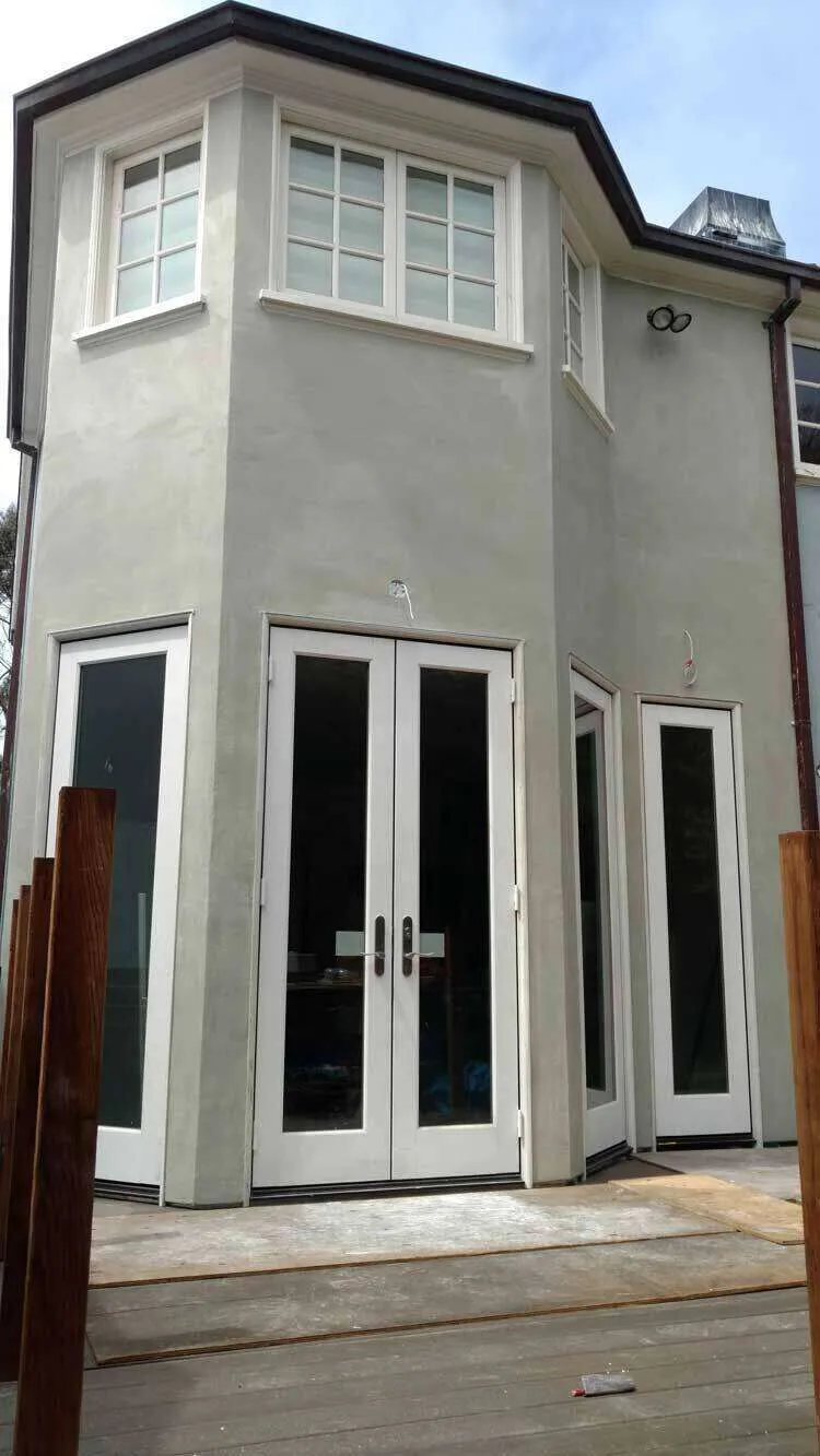 Corner of a house with smooth stucco finish, windows, and double doors leading to a deck.
