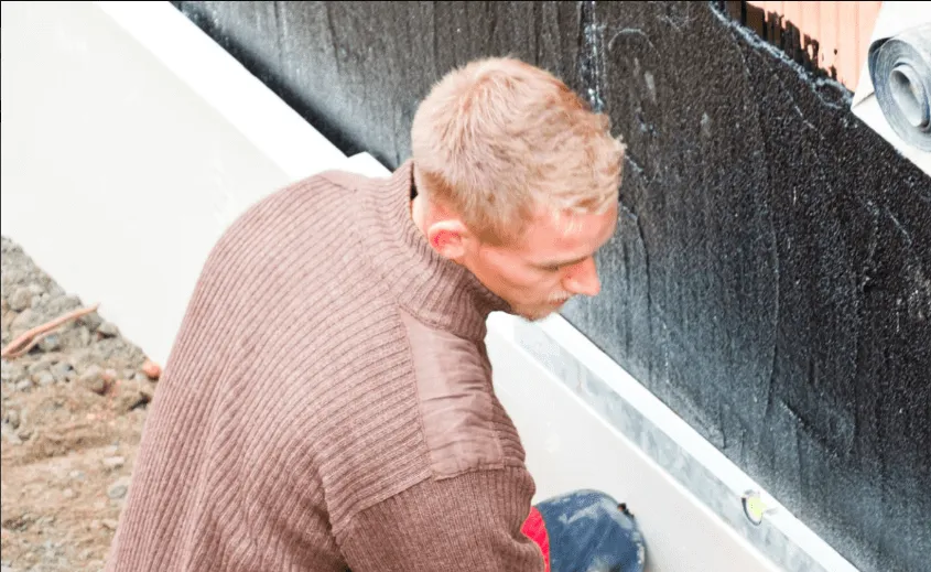 Person applying waterproofing membrane to a foundation wall with black material.