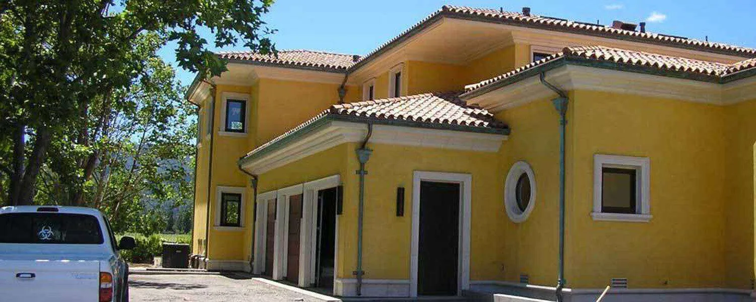 Large yellow stucco house with tiled roof and white truck in driveway, surrounded by trees.